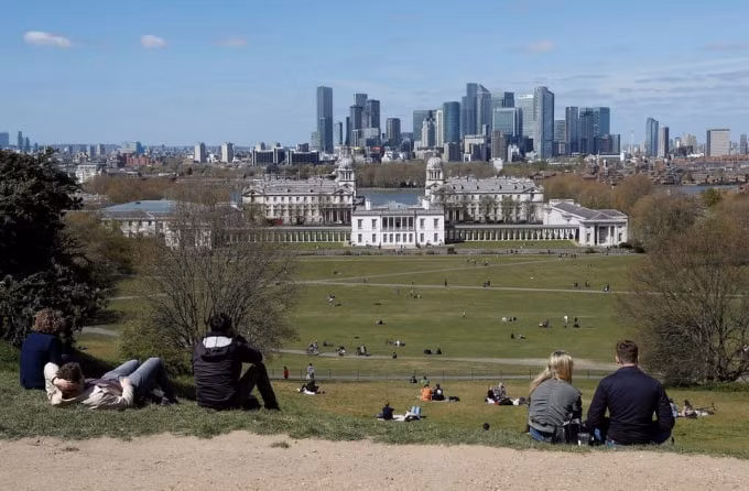 Du khách đến thăm Công viên Greenwich ngồi và nhìn về phía khu tài chính Canary Wharf khi các hạn chế về khóa cửa được nới lỏng trong bối cảnh đại dịch COVID-19 lây lan ở London, Anh, ngày 25 tháng 4 năm 2021. @ REUTERS
