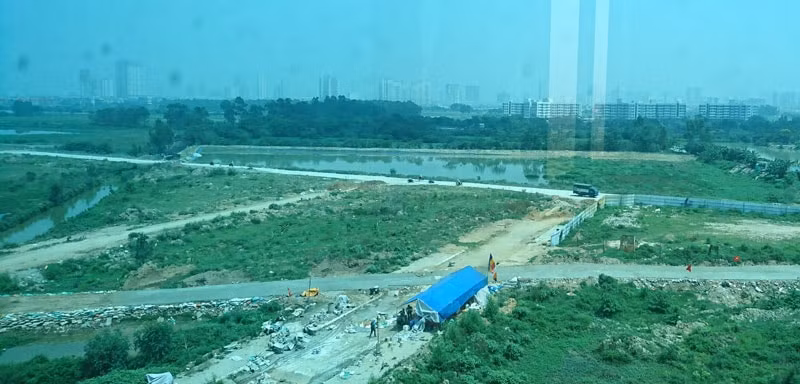 Đoạn giữa của 2 con đường. Đường do CĐT làm đang có xe tải và nhiều xe máy chạy ở xa góc ống kính. Đường do dân Bắc Lãm chiếm đất CĐT làm có lán xanh bên cạnh.